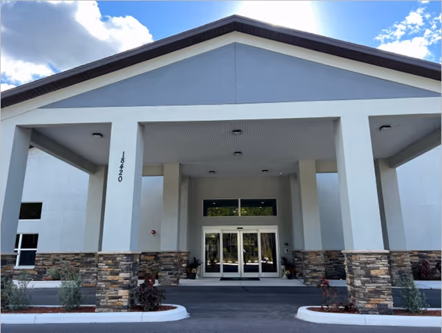 Front exterior view of a senior living facility building with a covered entrance supported by stone and white pillars, glass double doors, and the address number 18420 displayed on one pillar. The sky is partly cloudy with sunlight visible.