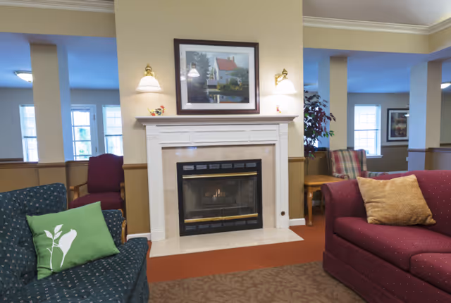 A cozy senior living facility common area featuring a lit fireplace with a white mantel, a framed painting above it, and two wall-mounted lamps on either side. The room has comfortable upholstered chairs and sofas in green and maroon colors with cushions, a side table with a plant, and large windows letting in natural light.