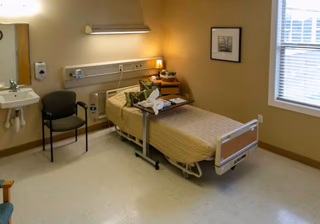 A simple, clean patient room with a single hospital bed covered in a beige bedspread, a small overbed table with a towel animal and some items on it, a chair next to a wall-mounted sink with soap dispenser, a small bedside table with a lamp and basket, a framed picture on the wall, and a window with blinds letting in natural light.