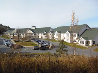 Exterior view of a large senior living facility building with green roofs and multiple windows, surrounded by a parking lot with several cars and some trees and shrubs in the foreground under a clear sky.