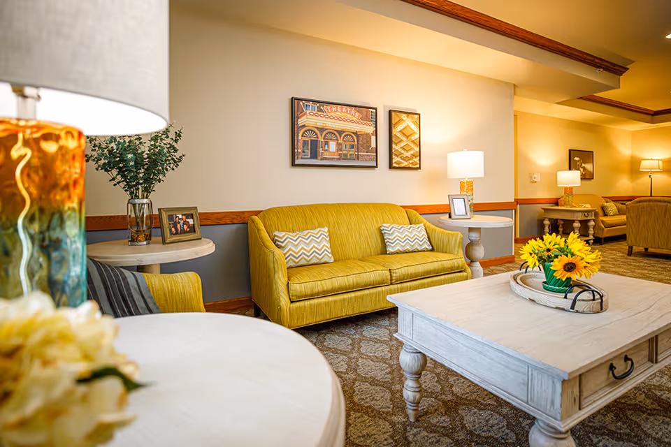 A cozy living room area with a yellow sofa and matching armchair, decorated with patterned pillows. There are two side tables with lamps and framed photos, a white wooden coffee table with a tray holding a vase of sunflowers, and framed artwork on the wall. The room has warm lighting and a patterned carpet.