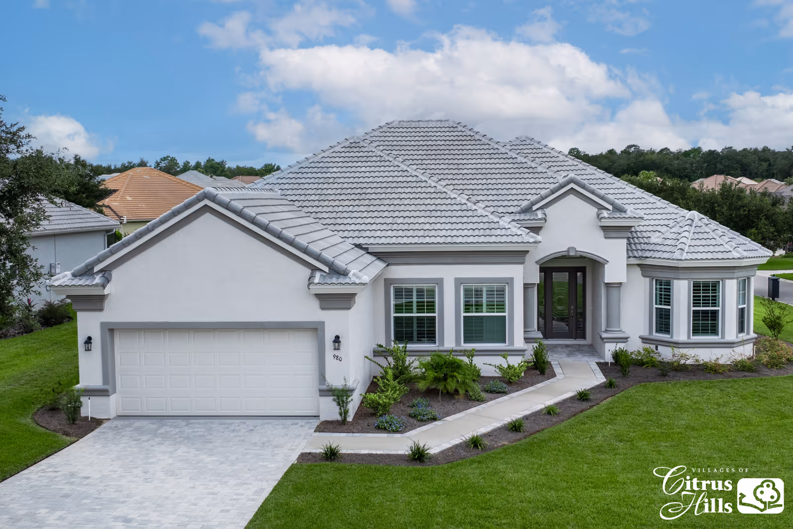 Front exterior view of a single-story house with a gray tiled roof, white walls, a two-car garage, and a well-maintained lawn and landscaping under a partly cloudy blue sky.