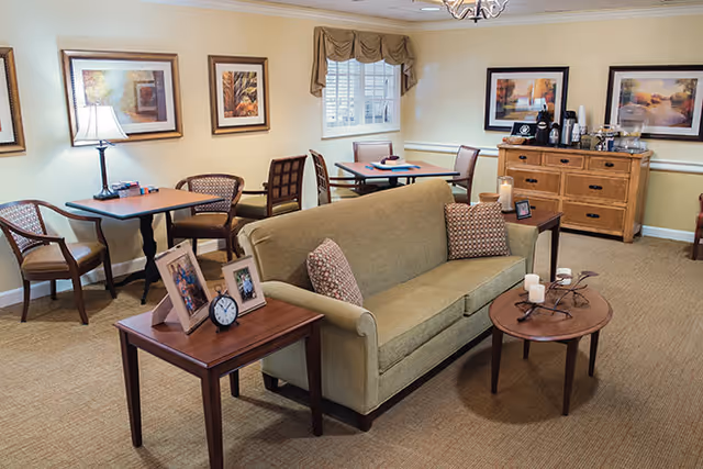 A comfortable communal lounge with a green sofa, side tables with framed photos, small dining tables and chairs, framed artwork, and a beverage station.