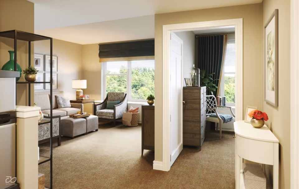 Bright furnished living room opening to a small bedroom area, with sofa and armchairs, a dresser, and large windows showing greenery outside.