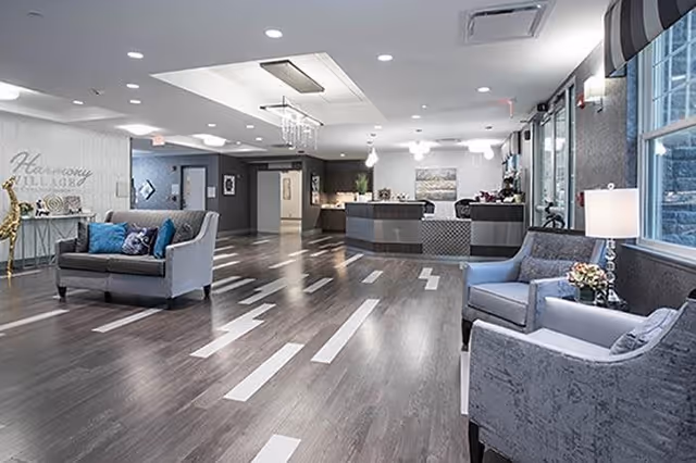 Spacious and modern senior living facility lobby with dark wood flooring featuring white rectangular inlays, comfortable gray sofas and armchairs with blue and patterned cushions, a reception desk in the background, decorative lighting fixtures on the ceiling, and large windows allowing natural light to brighten the space.