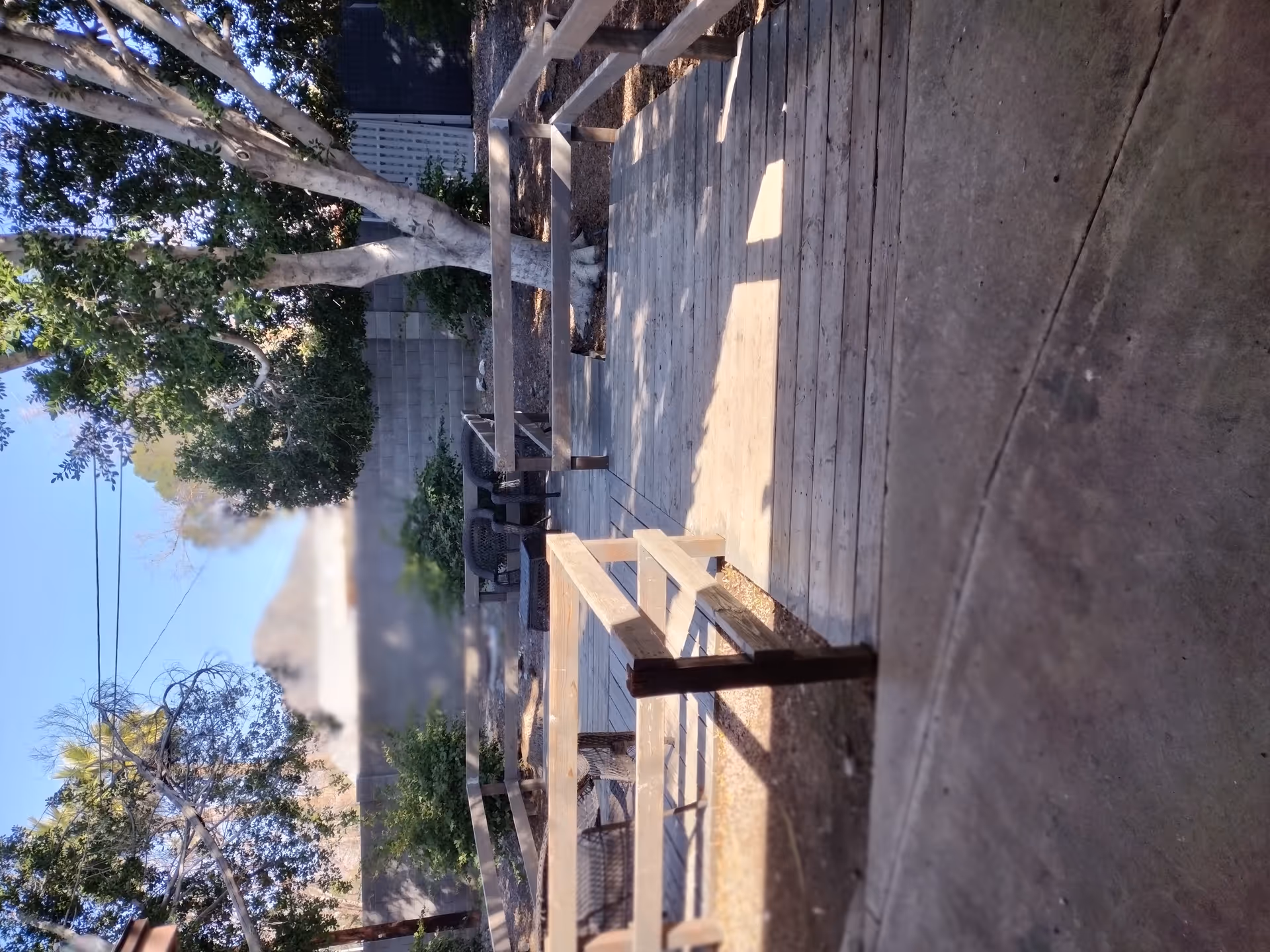Outdoor patio area with wooden decking and railing, surrounded by trees and shrubs. There are some chairs and a table in the background, with a concrete pathway adjacent to the deck. The scene is sunlit with shadows cast by the trees.