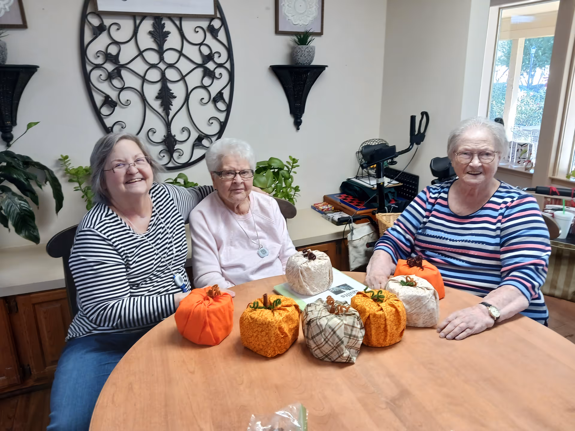 Three elderly women sitting around a wooden table with fabric pumpkins arranged on it, smiling and posing for the photo in a cozy room with plants and decorative wall art.