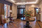 A warmly lit interior entryway featuring a decorative black front door with glass panels. The room has a round wooden table with a large floral arrangement in the center. Two beige upholstered chairs with a small round side table and lamp are positioned near a window with curtains. The floor is wooden with a patterned design, and there is a mirror and framed artwork on the walls.