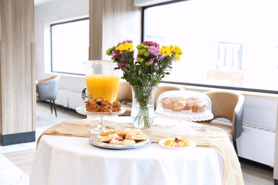 A round table covered with a white tablecloth and a beige runner, displaying a glass dispenser filled with orange juice, a vase with colorful flowers, a plate of pastries, a covered plate of muffins, and a small plate of fruit. The background shows a bright room with large windows and beige chairs.