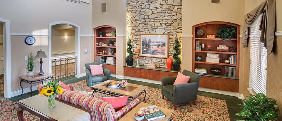 A cozy living room area with a striped sofa and two green armchairs, each with pink cushions. There is a wooden coffee table with books and a flower vase on it. The room features a stone fireplace with decorative plants and a framed landscape painting above it. Built-in wooden shelves with books and decorative items flank the fireplace. A large window with blinds and curtains lets in natural light, and a side table with a lamp and a clock on the wall are visible.
