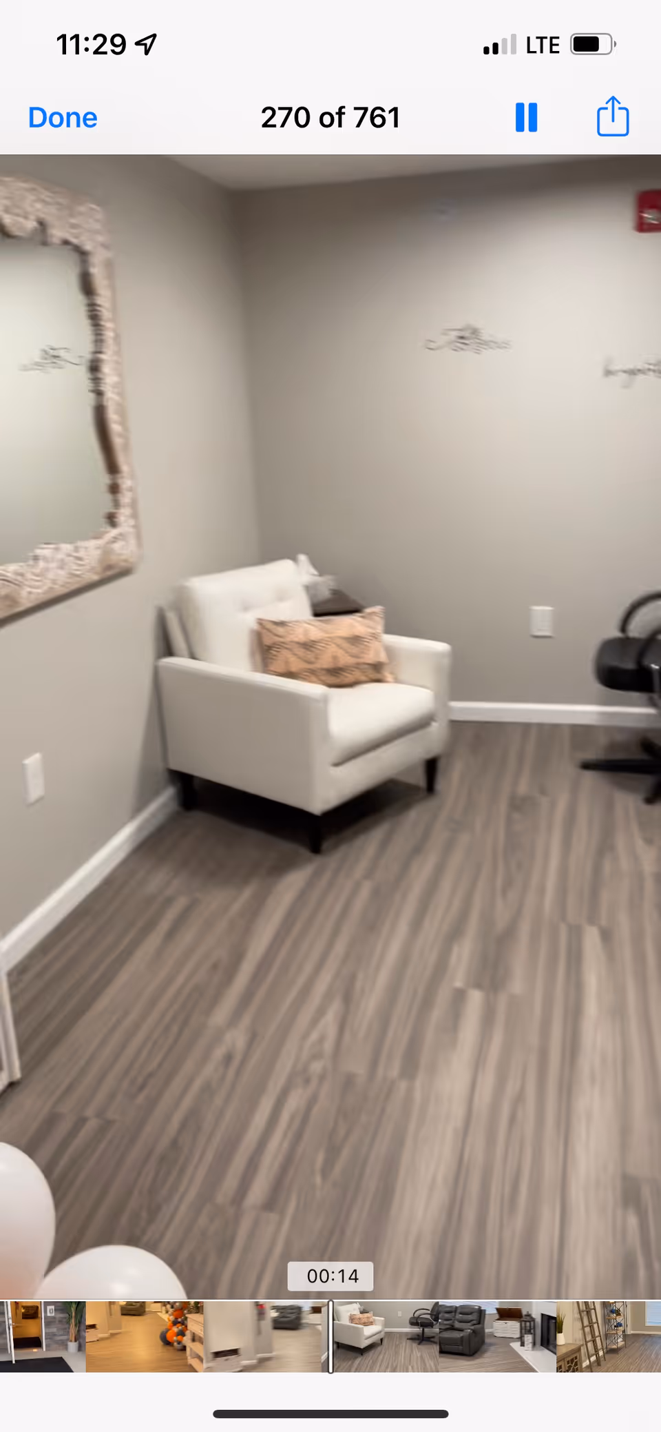 A cozy interior room with light gray walls and wood-patterned flooring. There is a white armchair with a decorative pillow placed against the wall, a large ornate mirror hanging on the left wall, and a black office chair partially visible on the right side.