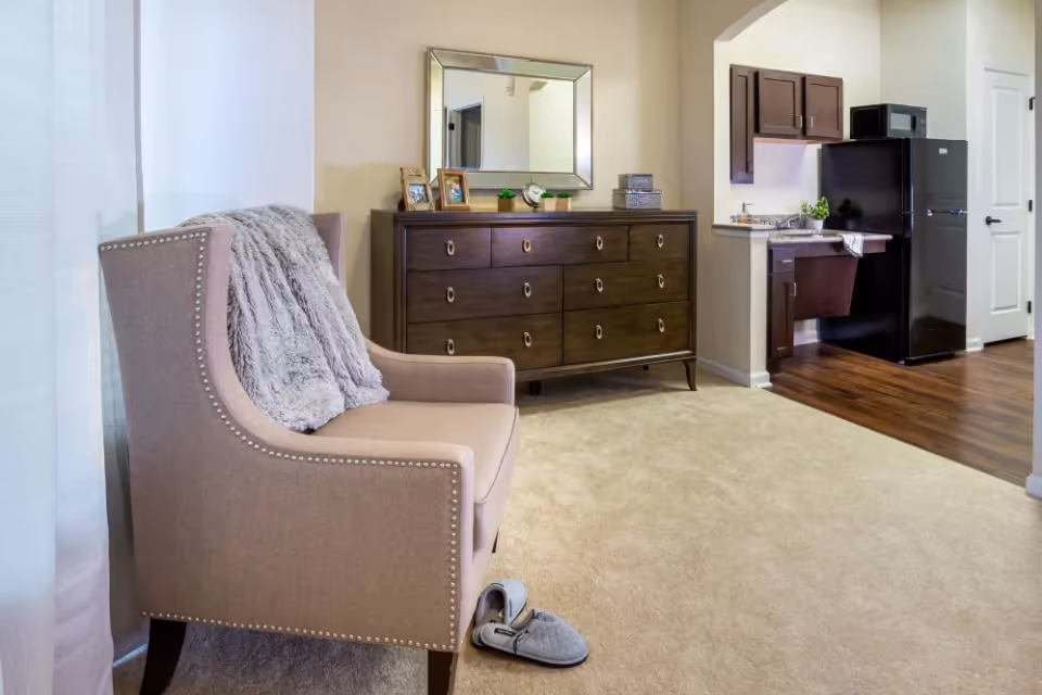 A cozy interior space featuring a beige upholstered armchair with a gray throw draped over it, a dark wooden dresser with a large mirror above it, and a small kitchen area with dark cabinets, a black refrigerator, and a microwave. The floor is carpeted in the sitting area and transitions to wood flooring in the kitchen.