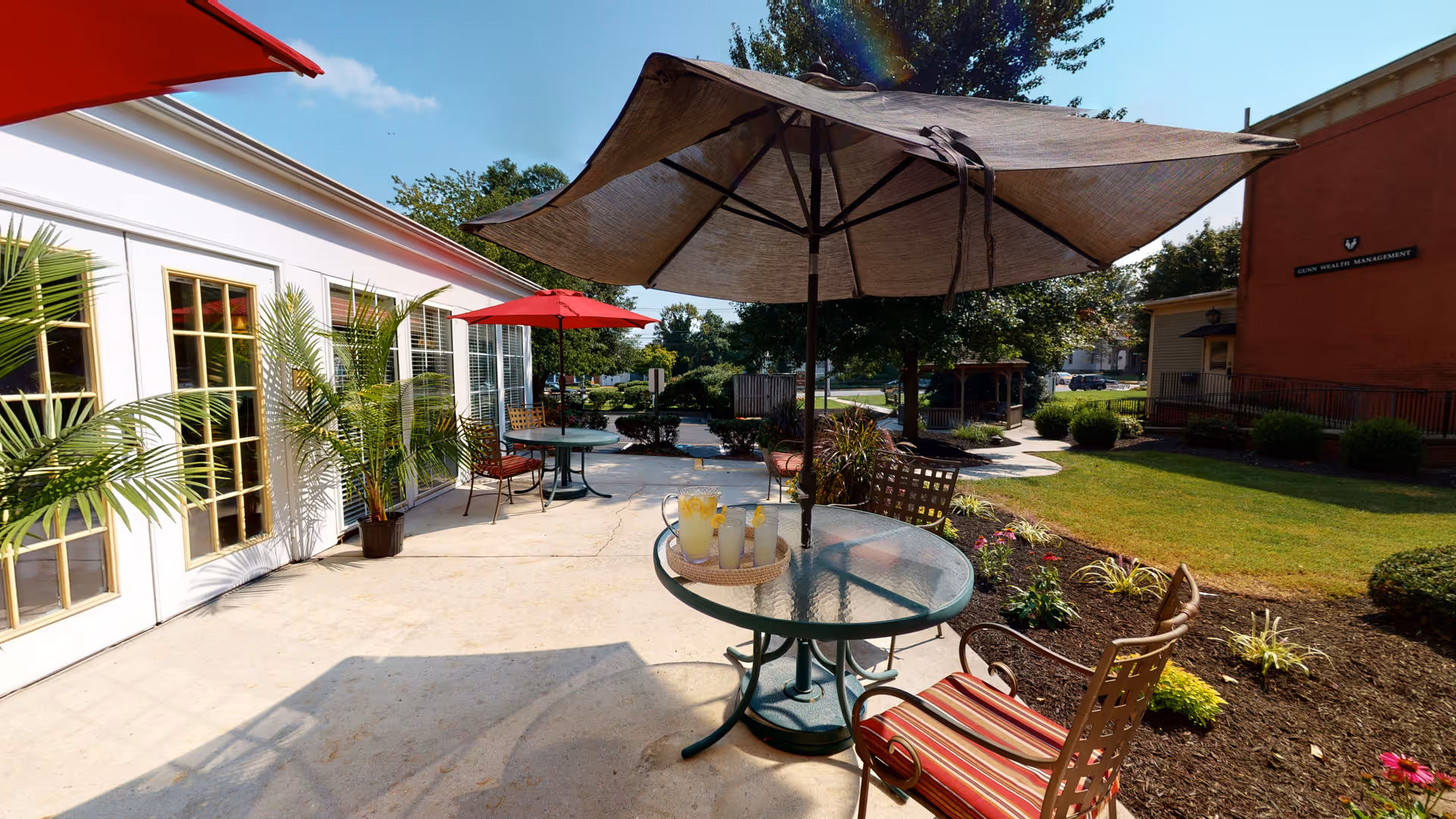 Outdoor patio with glass tables and umbrellas, potted plants, and lawn beside a building.