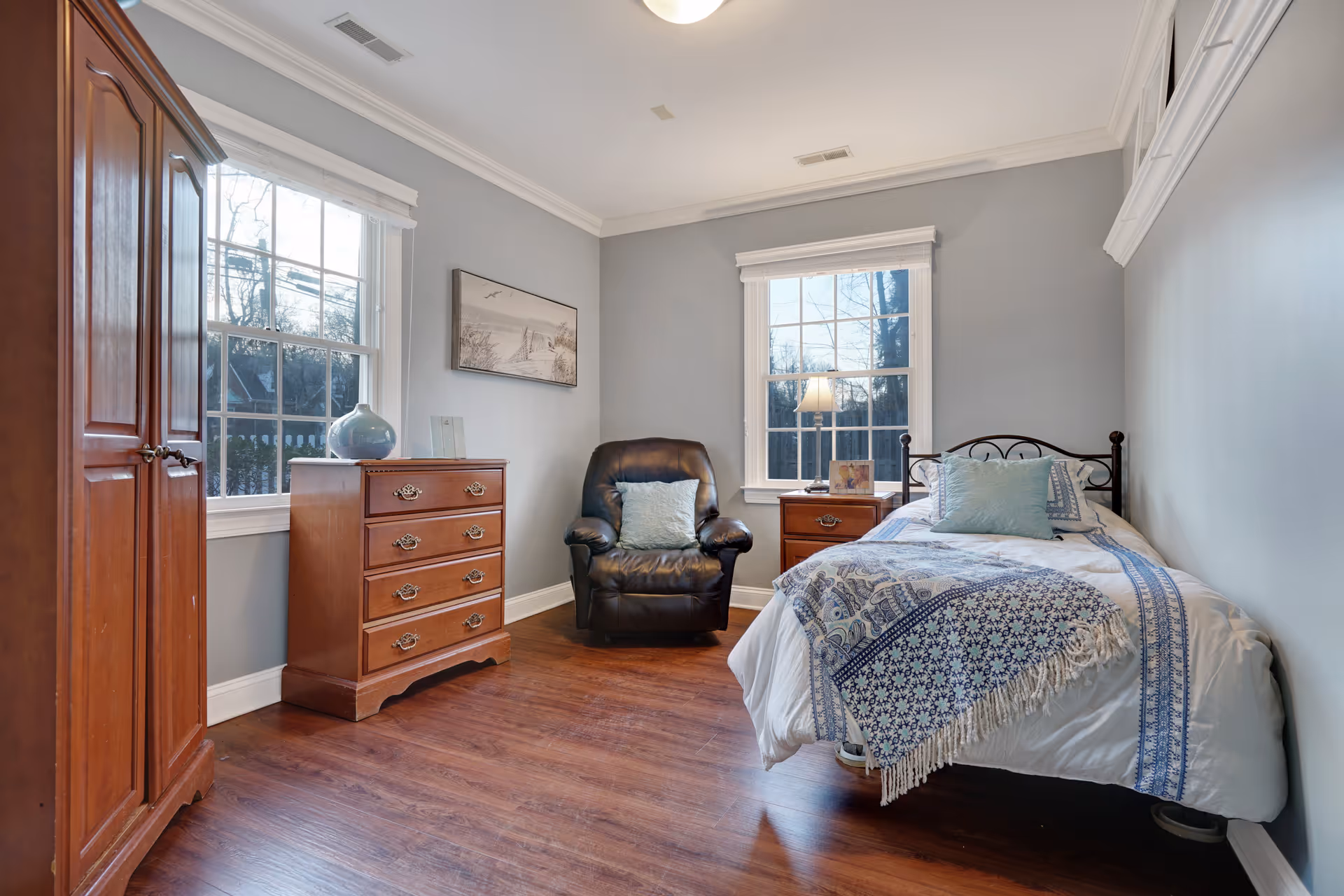 A cozy bedroom with light gray walls and wooden flooring. The room features a single bed with white and blue patterned bedding, a wooden nightstand with a lamp and a framed photo, a black leather armchair with a light blue pillow, a wooden chest of drawers with a decorative vase, and a wooden wardrobe. Two large windows allow natural light to fill the room.