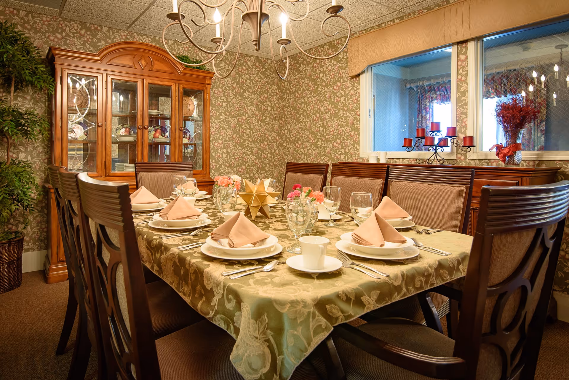 A formal dining room set for a meal with a long table covered by a patterned tablecloth, folded napkins, place settings, wooden chairs, a china cabinet and floral wallpaper.