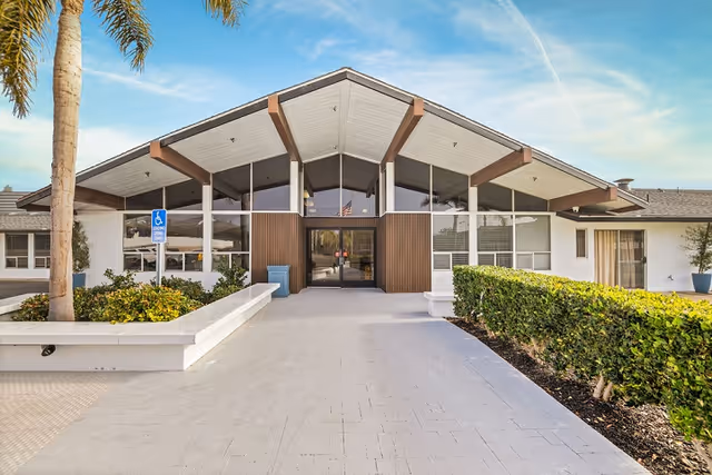 Front exterior view of Capistrano Beach Care Center building with a wide walkway leading to glass entrance doors, surrounded by greenery and palm trees under a blue sky.