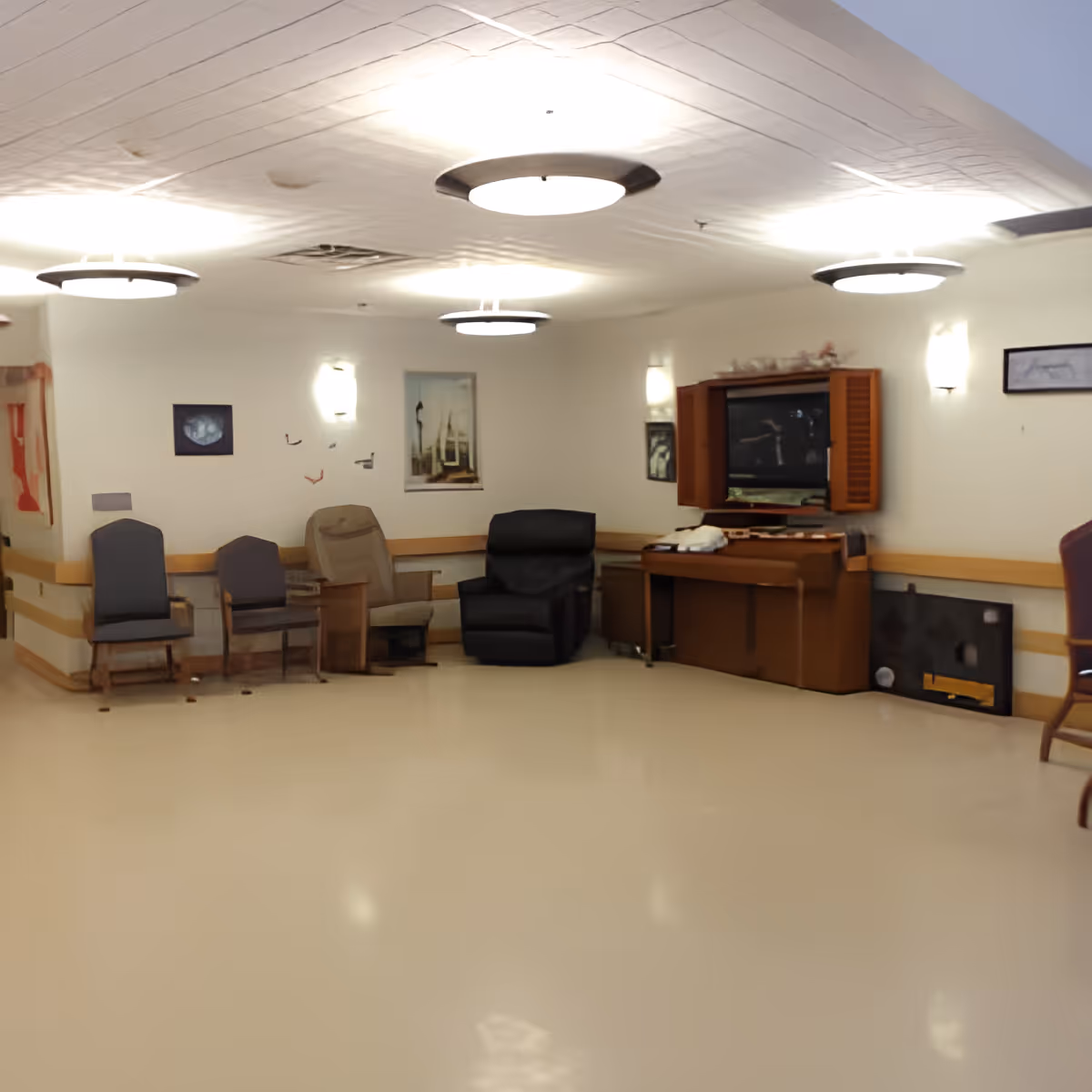 A common area in Orchard Manor featuring several chairs arranged along the walls, a television set inside a wooden cabinet, wall-mounted lights, and framed pictures on the walls. The room has a smooth, light-colored floor and ceiling lights.