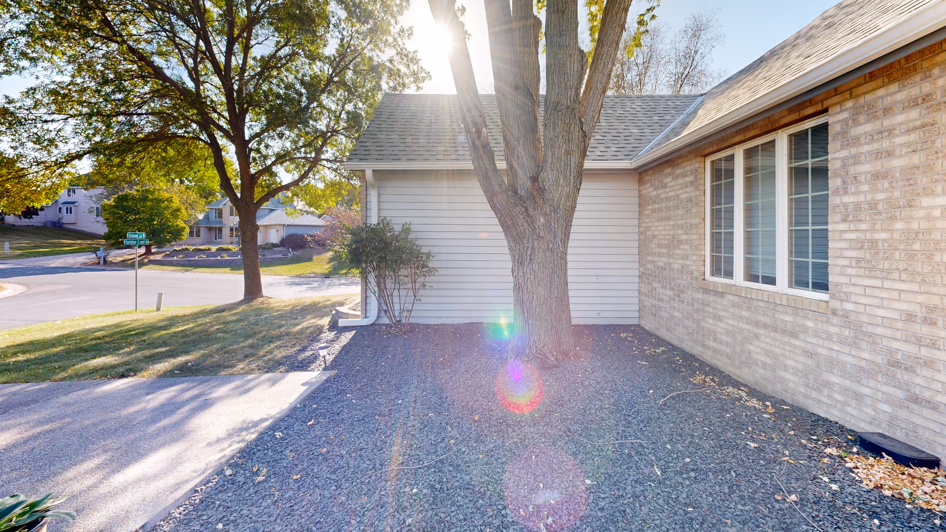 Sunlit exterior side of a single-story brick and siding house with a tree near the foundation and a street in the background.