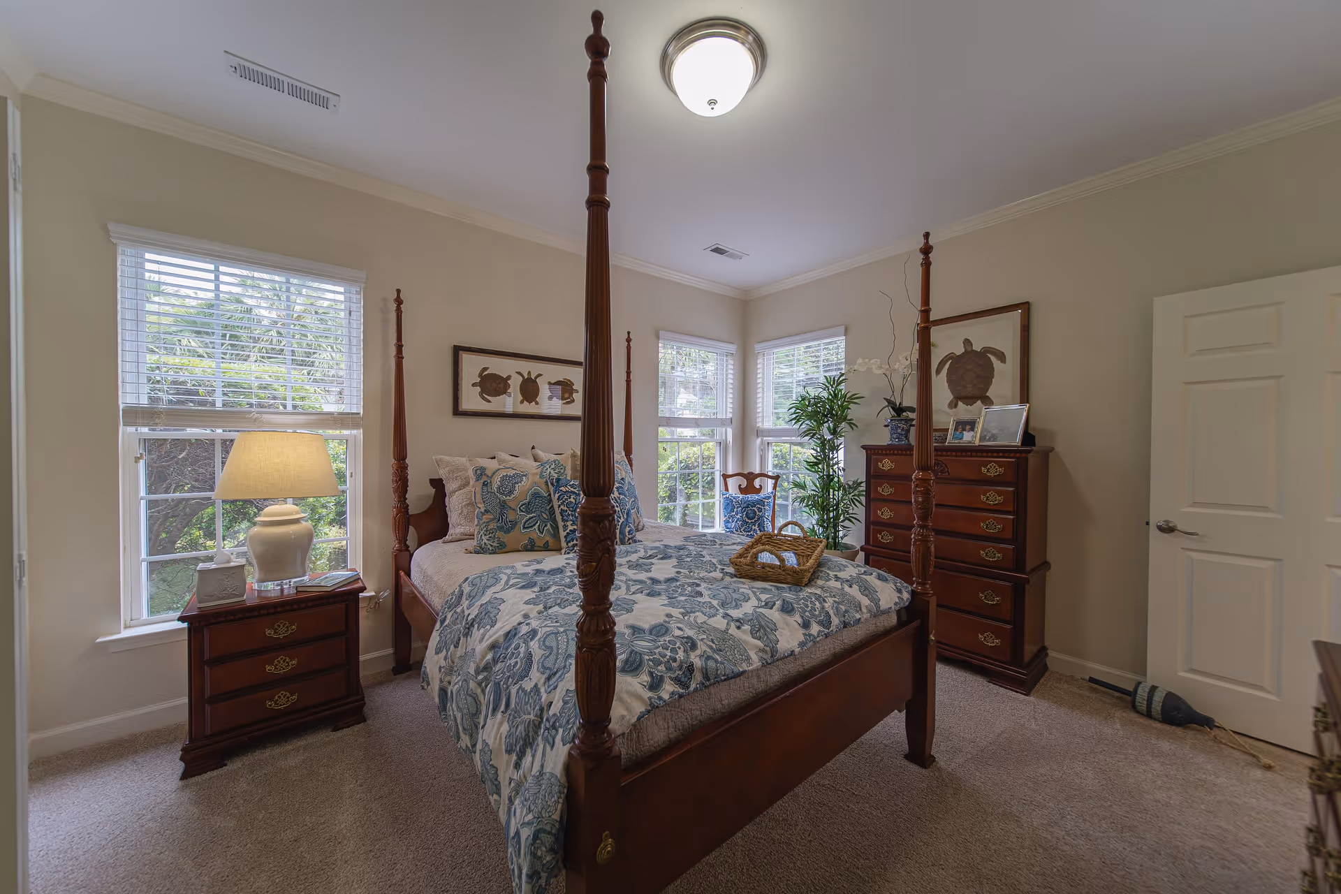 A cozy bedroom with a wooden four-poster bed adorned with blue and white floral bedding and multiple pillows. There are two windows with white blinds letting in natural light. A wooden nightstand with a lamp and tissue box is beside the bed. A wooden dresser with framed pictures and a plant on top is against the wall. A wooden chair with a blue cushion and a tall green plant are near the corner windows. The walls are painted beige and the carpet is light-colored.