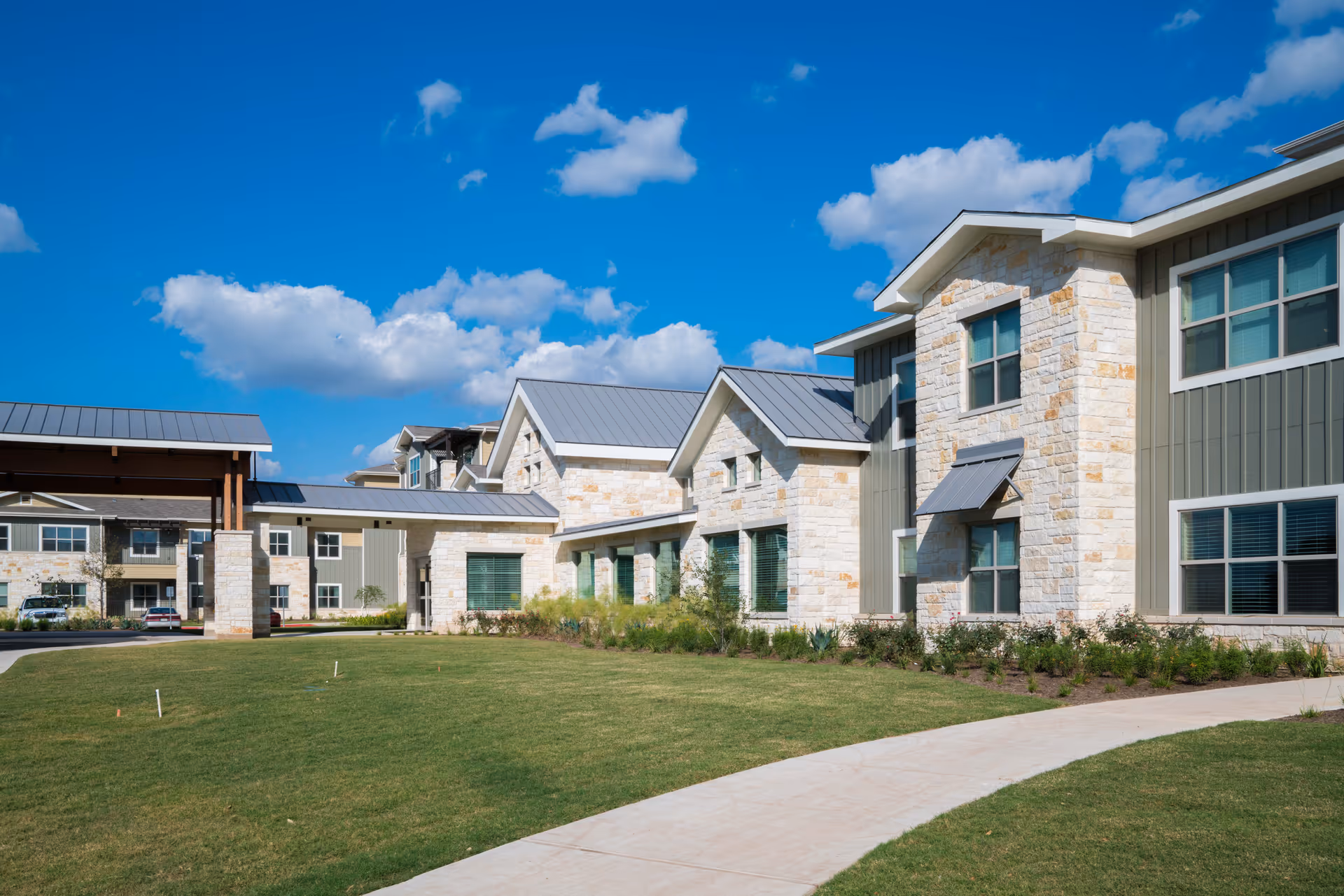Exterior view of The Delaney at Georgetown Village senior living facility showing a modern building with stone and siding facade, multiple windows, a covered entrance, and a well-maintained lawn under a blue sky with scattered clouds.