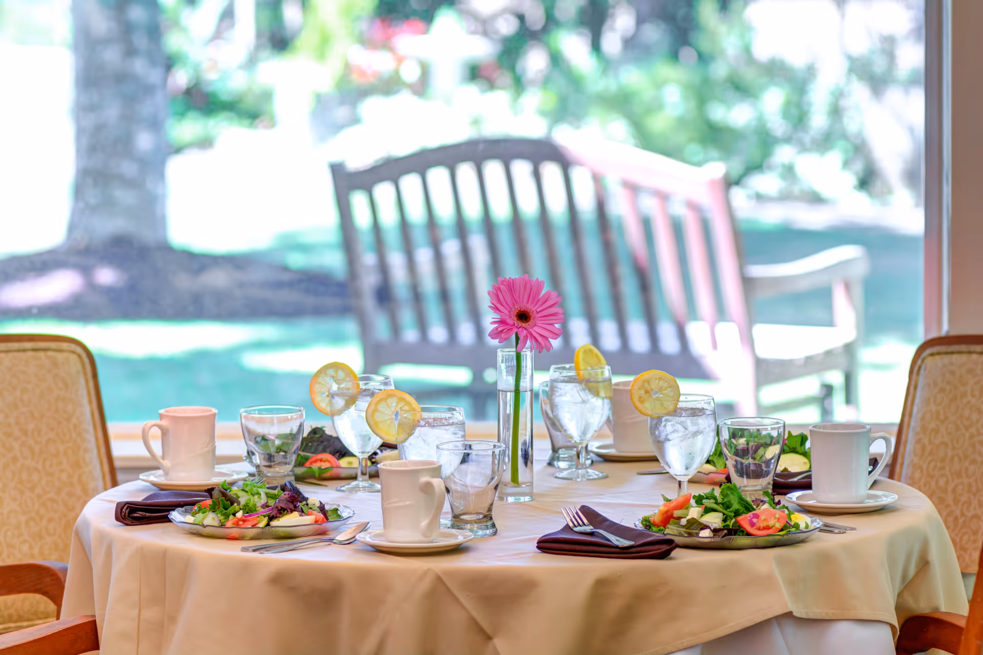 A round dining table set for four with plates of salad, glasses of water with lemon slices, coffee cups, and silverware. A single pink flower in a vase is placed in the center of the table. The background shows a window with an outdoor view of a wooden bench and greenery.