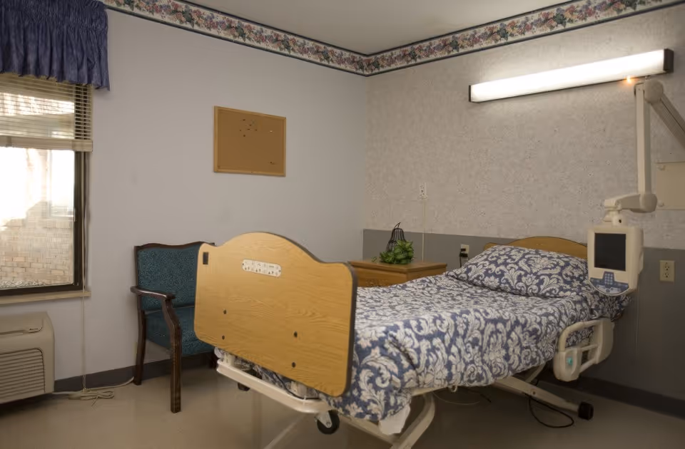 A single adjustable hospital-style bed with blue patterned bedding in a small assisted living resident room with a chair, nightstand and window.