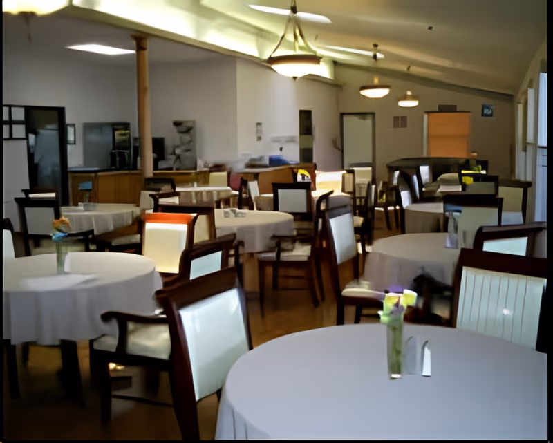 A dining room with multiple round tables covered with white tablecloths, each surrounded by wooden chairs with white cushioned backs and seats. Small flower vases are placed on the tables. The room has a high ceiling with hanging light fixtures and a wooden floor.