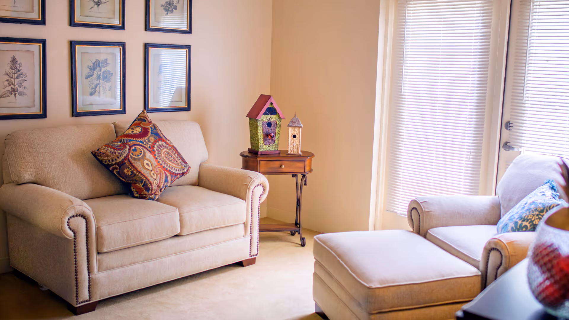 A cozy living room area with a beige loveseat and armchair, each adorned with decorative pillows. A small wooden side table between the seats holds two colorful birdhouses. The room has framed botanical prints on the wall and a window with closed blinds letting in soft natural light.