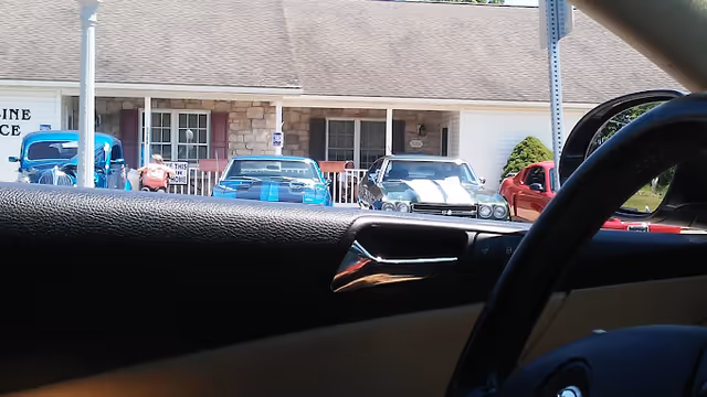 View from inside a car of the front porch of a single-story senior living building with vintage cars parked in front and a person seated on the porch.