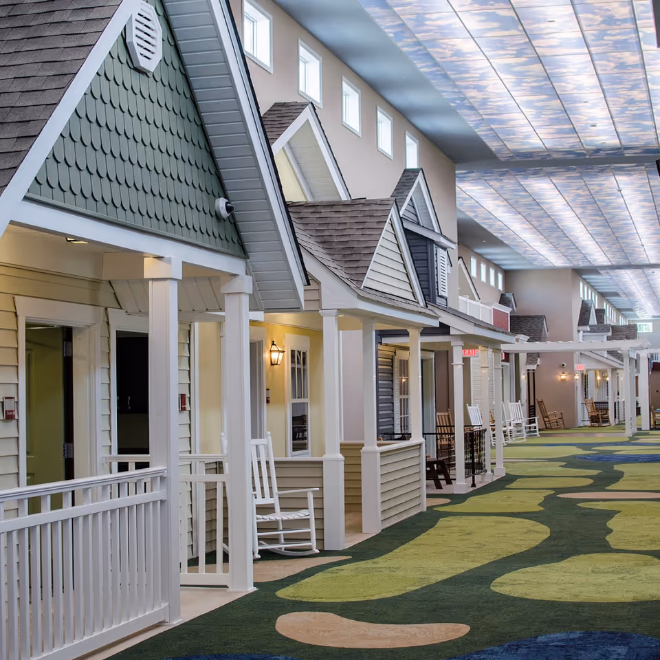 Indoor corridor of a senior living facility styled as a row of small house-fronts with porches and rocking chairs beneath a skylight ceiling.
