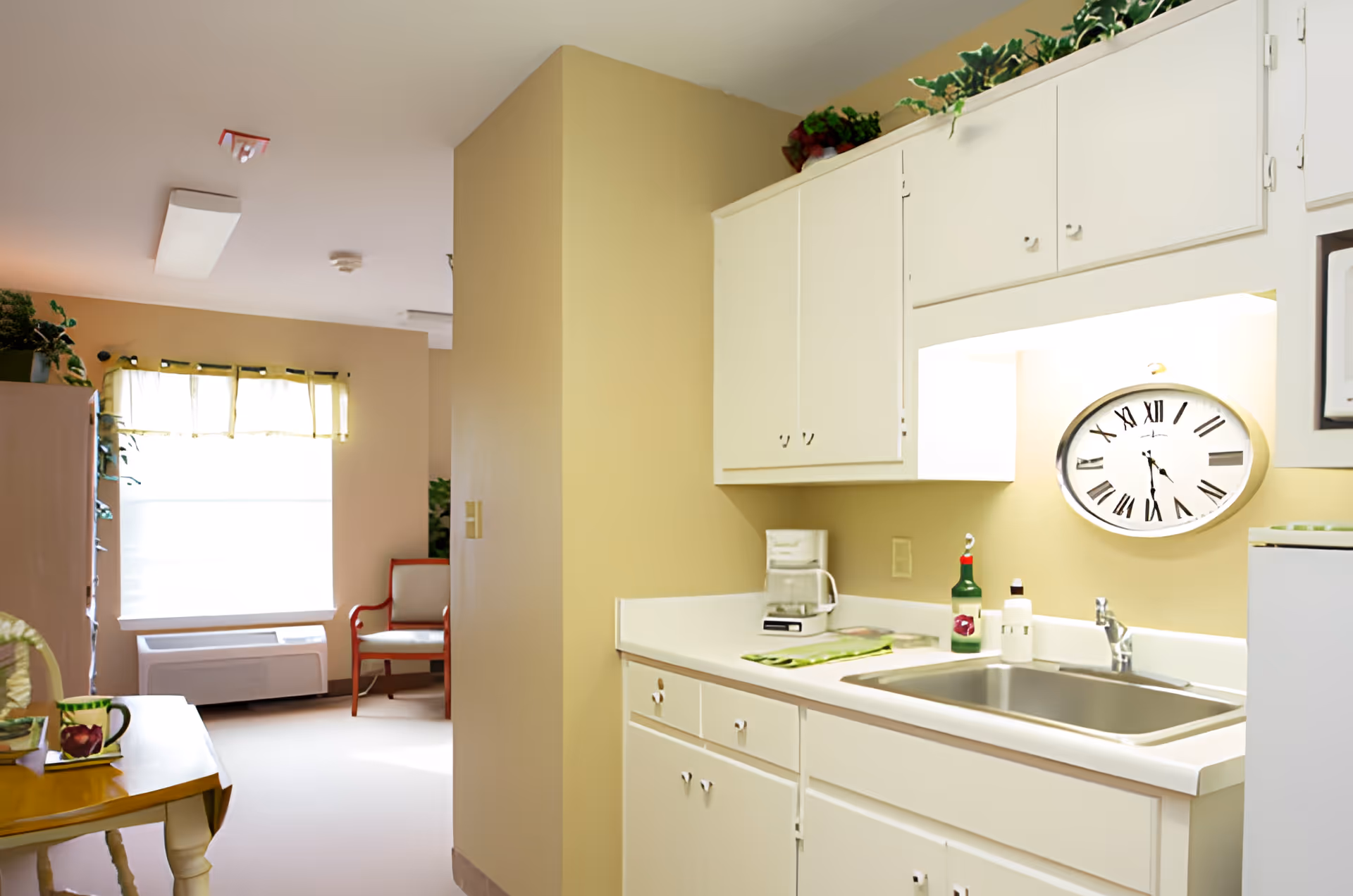 A small kitchen area with white cabinets, a sink, a coffee maker, and a wall clock with Roman numerals. The kitchen opens into a room with a window covered by a light curtain, a red and white chair, and a wooden table with a cup and a plant on it.