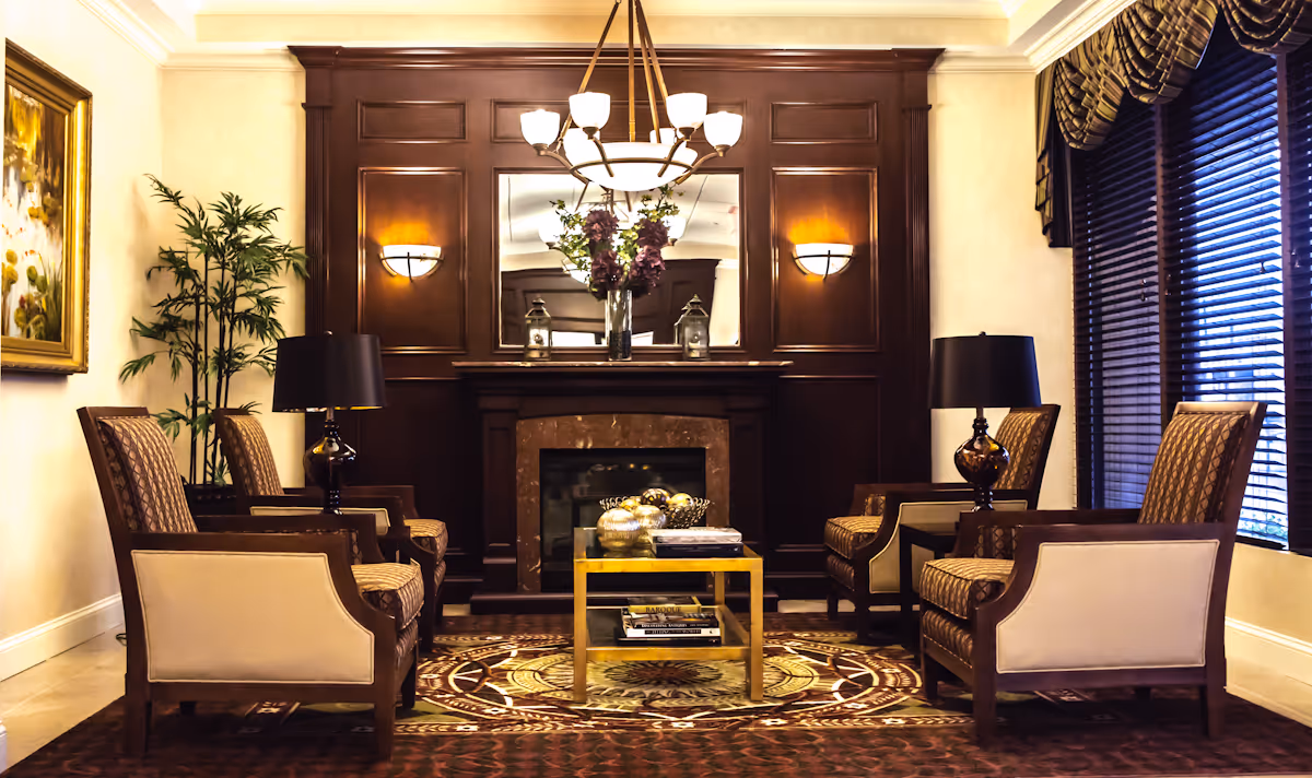 A cozy living room with four upholstered armchairs arranged around a small glass coffee table on a patterned rug. The room features a dark wood-paneled wall with a fireplace, a large mirror above it, two wall sconces, and a chandelier hanging from the ceiling. There are two table lamps on side tables next to the chairs, a tall potted plant in the corner, a large window with blinds and valances, and a framed painting on the left wall.