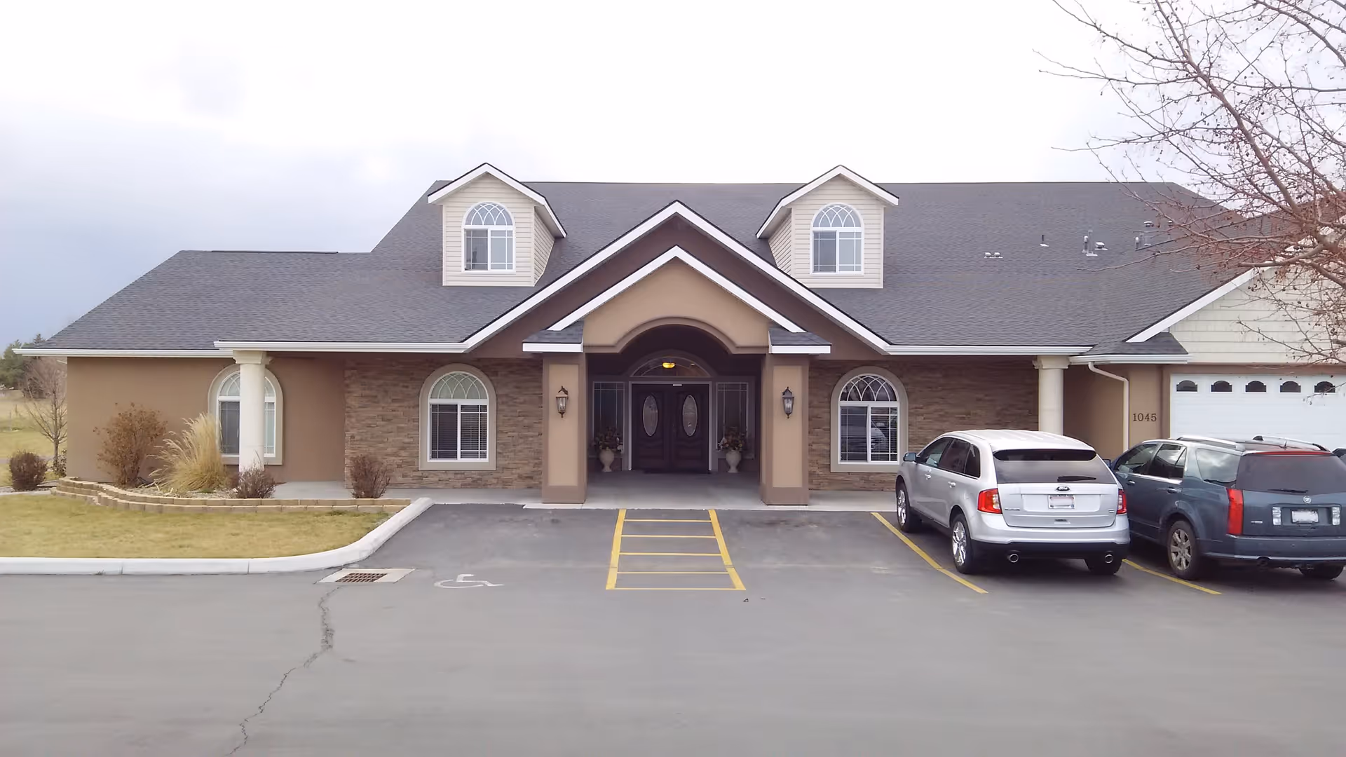 Front exterior view of a single-story assisted living facility building with a peaked roof and dormer windows. The entrance has double doors under a covered porch supported by columns. There are three cars parked in front of the building and some landscaping with grass, bushes, and a tree without leaves.