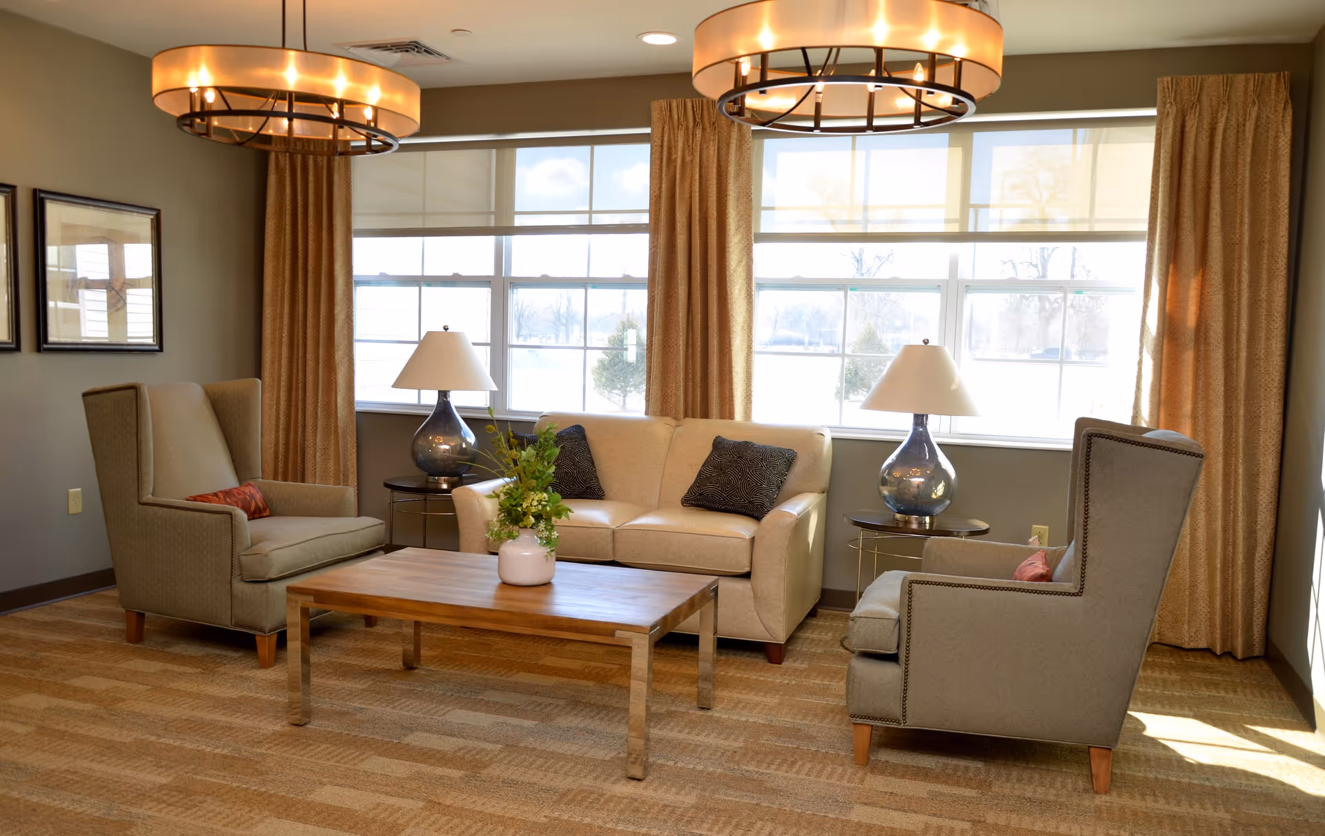 A cozy living room area with a beige sofa, two armchairs, a wooden coffee table with a vase of flowers, two table lamps on side tables, large windows with beige curtains, and two round ceiling lights.