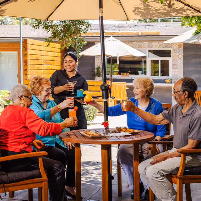 Four elderly people sitting around a wooden table outdoors under a large umbrella, raising their drinks in a toast. A server stands nearby holding a tray with additional drinks. The setting appears to be a patio area with a bar in the background.