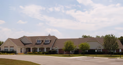 Single-story brick and stone community building with dormer windows, a curved driveway, parking spaces, and landscaped lawn under a partly cloudy sky.