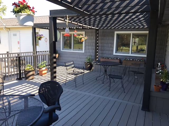 Outdoor patio area with a pergola casting striped shadows on the wooden deck. Several metal tables and chairs are arranged around the space. Various potted plants and hanging flower baskets decorate the area. The exterior wall of the building with windows is visible in the background.