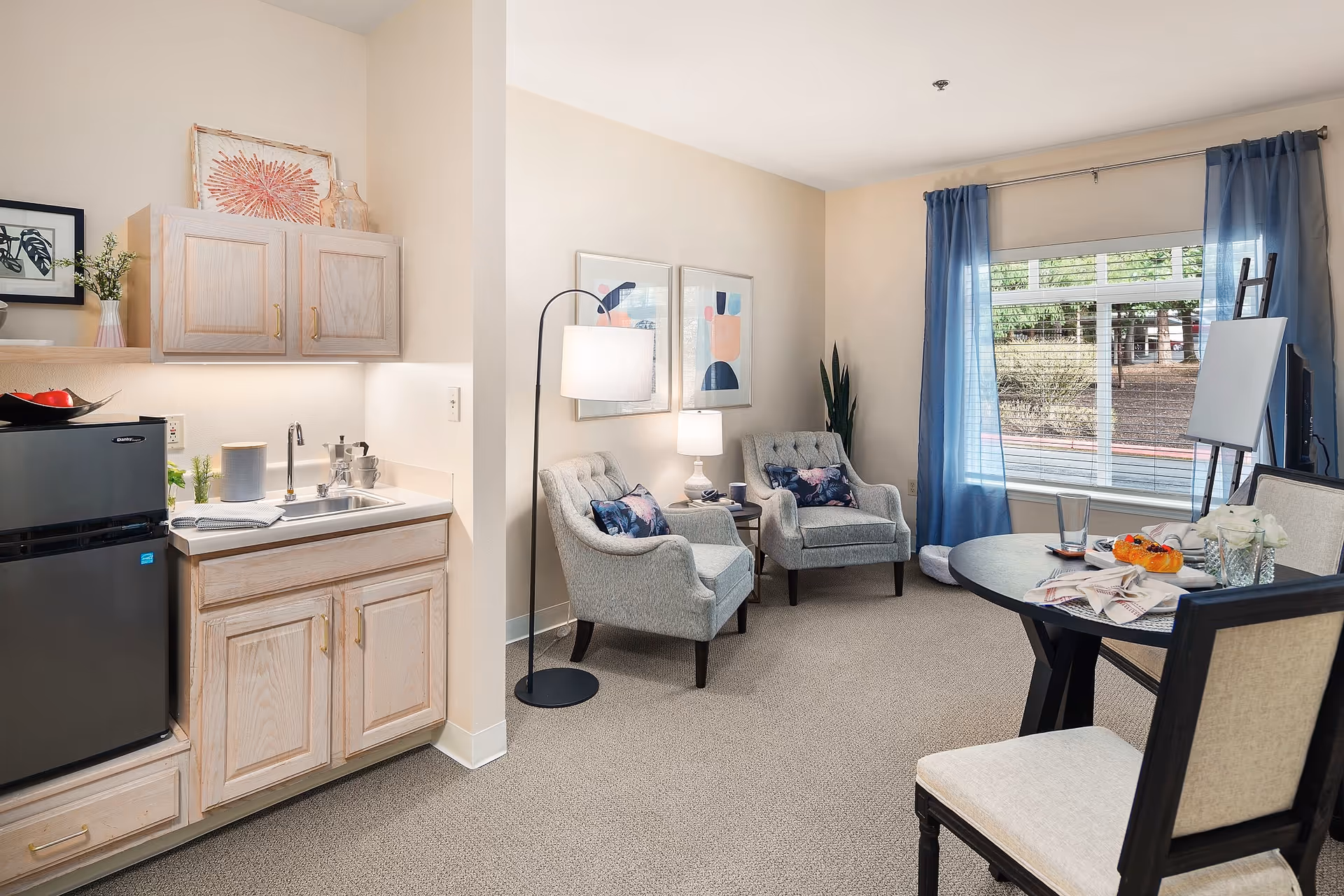 A cozy interior room featuring a small kitchenette with a black mini fridge, sink, and light wood cabinets. Adjacent to the kitchenette are two upholstered armchairs with decorative pillows, a floor lamp, and a side table with a lamp. A round dining table with chairs is positioned near a large window with blue curtains, allowing natural light to brighten the space. The room is decorated with framed abstract art and plants.
