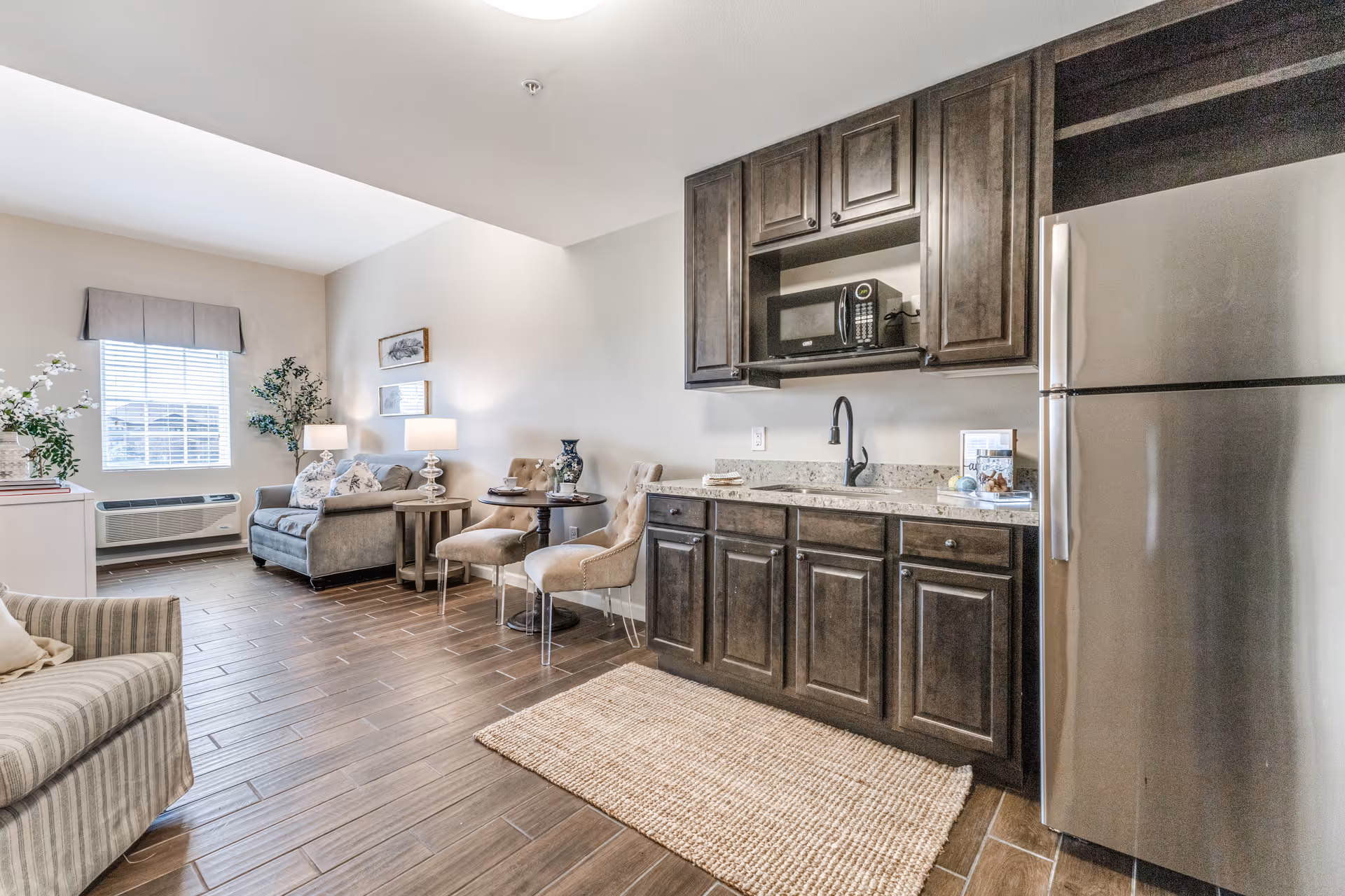 A bright and cozy senior living apartment interior featuring a small kitchenette with dark wood cabinets, a stainless steel refrigerator, a microwave, and a sink. Adjacent to the kitchenette is a small dining area with a round table and two upholstered chairs. The living area includes a gray loveseat, a side table with a lamp, and a window with a valance letting in natural light. The floor is covered with wood-look tiles and a woven rug in front of the kitchenette.