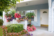 Front porch of a light blue house with white trim, decorated with hanging baskets and flower beds filled with colorful flowers, including pink, red, and purple blooms. The porch has white railings and a white door with a small window.