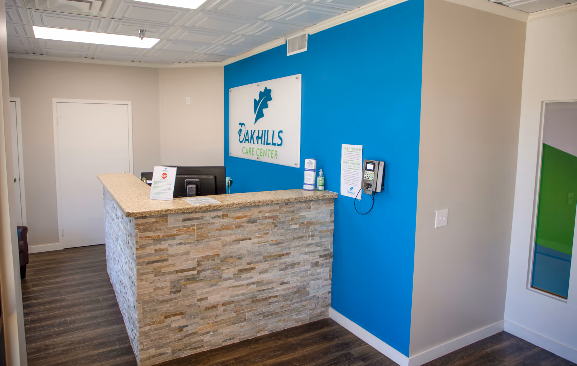 Reception desk area inside Oak Hills Care Center Skilled Nursing & Rehabilitation with a stone facade counter, a blue accent wall behind it featuring the Oak Hills Care Center logo, and a keypad entry system mounted on the wall.
