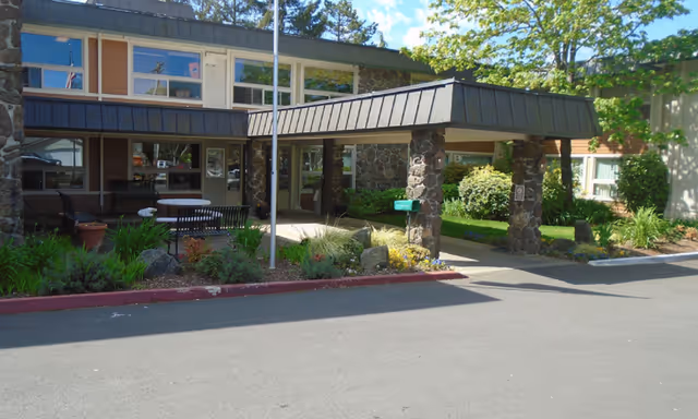 Entrance of South Hills Rehabilitation Center featuring a covered drop-off area supported by stone pillars, surrounded by landscaped greenery and outdoor seating with tables and chairs.