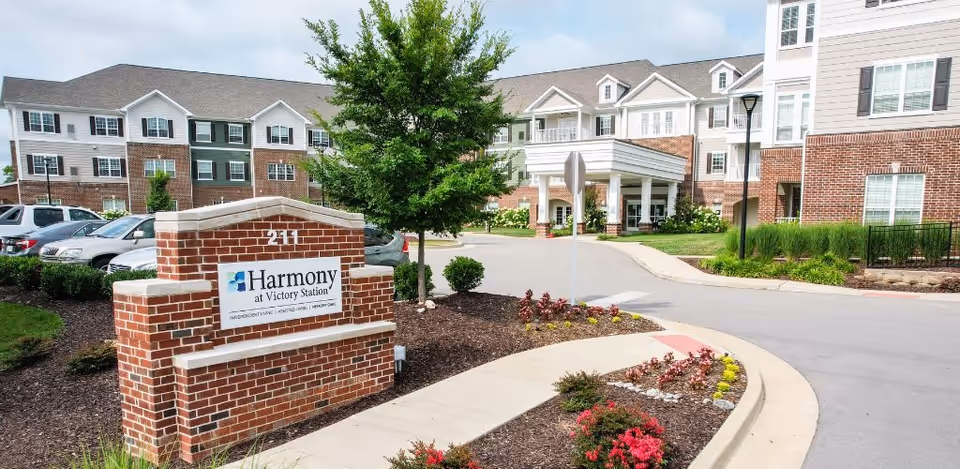 Exterior view of Harmony at Victory Station senior living facility showing a brick sign with the facility name, a driveway, landscaped flower beds, and a multi-story building with white siding and brick accents under a partly cloudy sky.