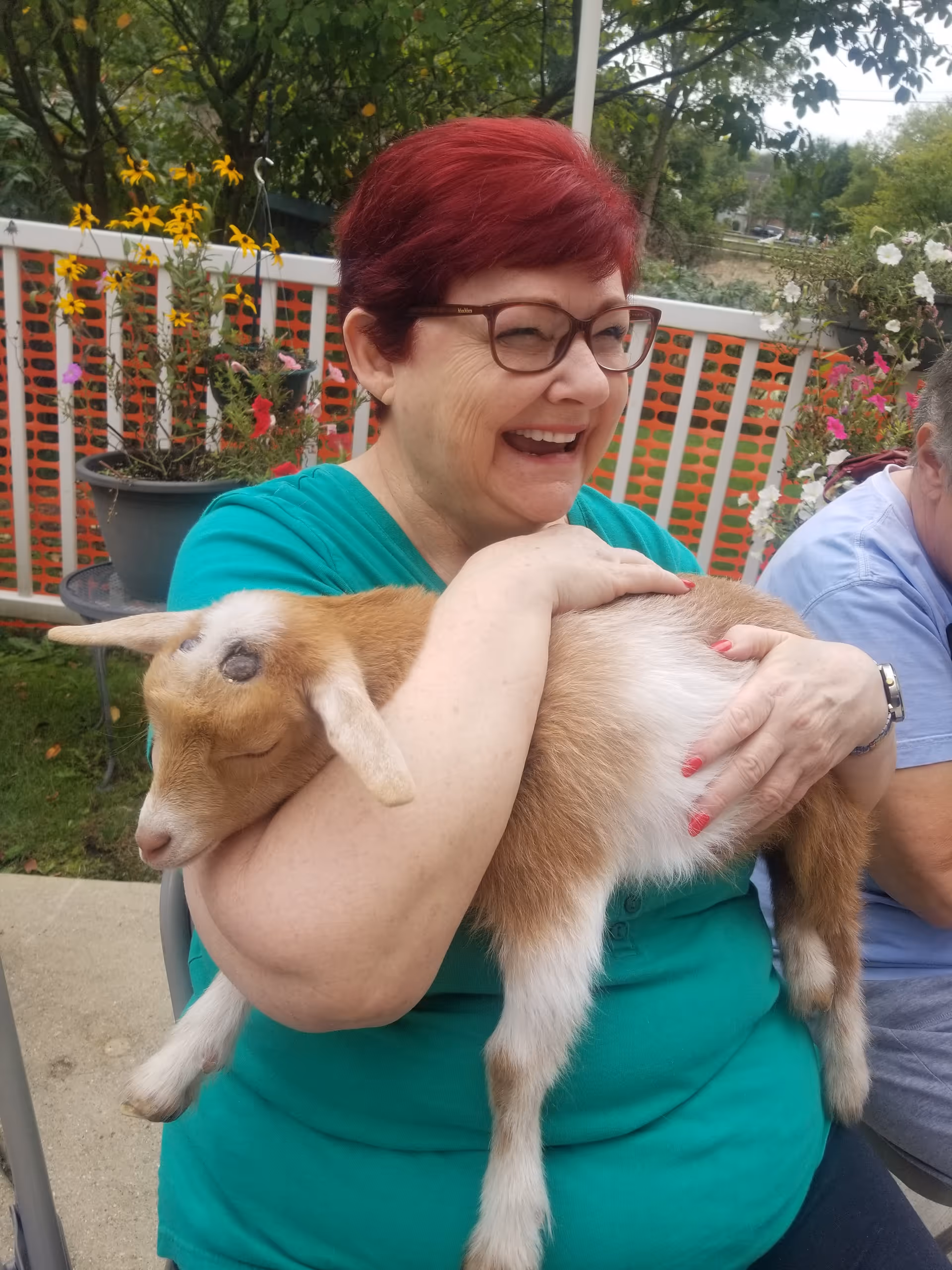 A smiling woman with short red hair and glasses wearing a green shirt is sitting outdoors holding a small brown and white goat in her arms. Behind her, there are potted flowers and a white fence with orange netting. Another person is partially visible sitting next to her.