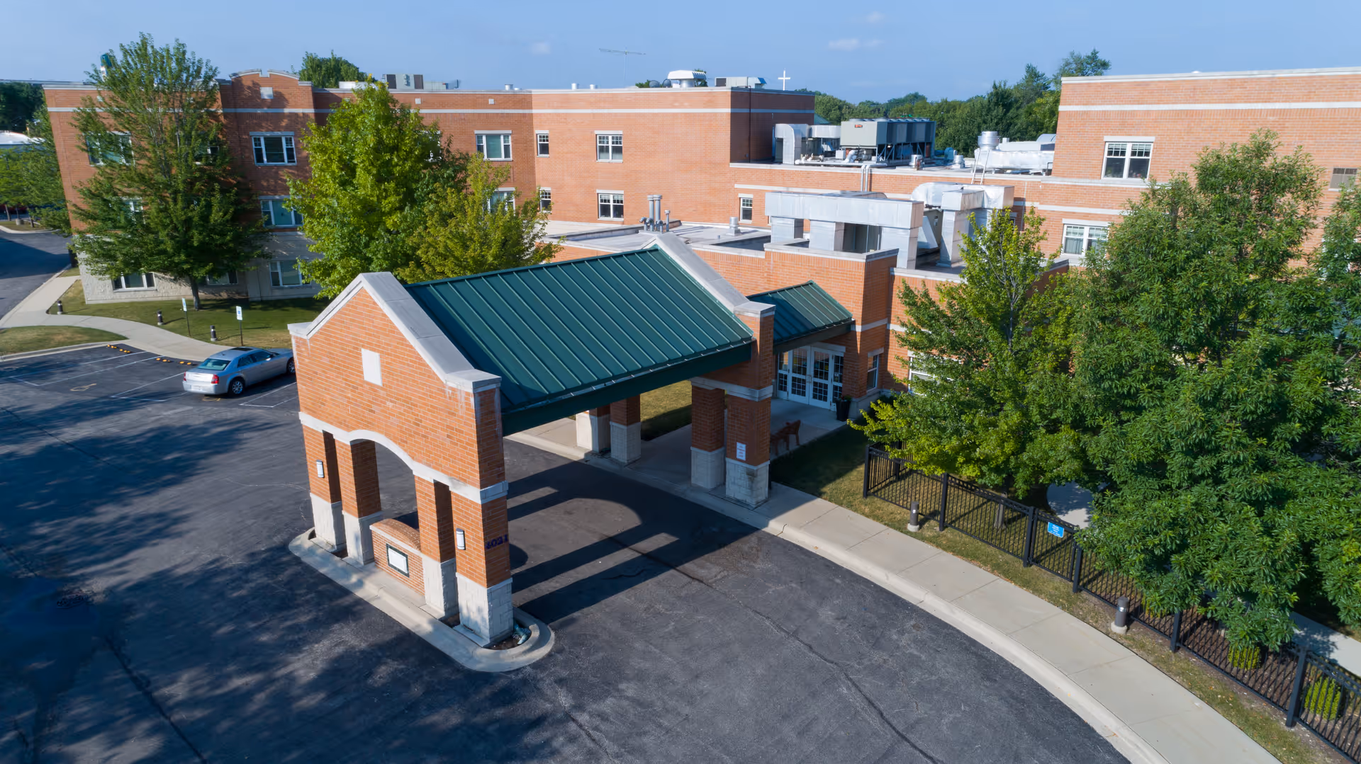 Exterior view of St. Joseph Village of Chicago showing a brick building with multiple windows, a covered entrance with a green roof, surrounding trees, a parking area, and a clear blue sky.