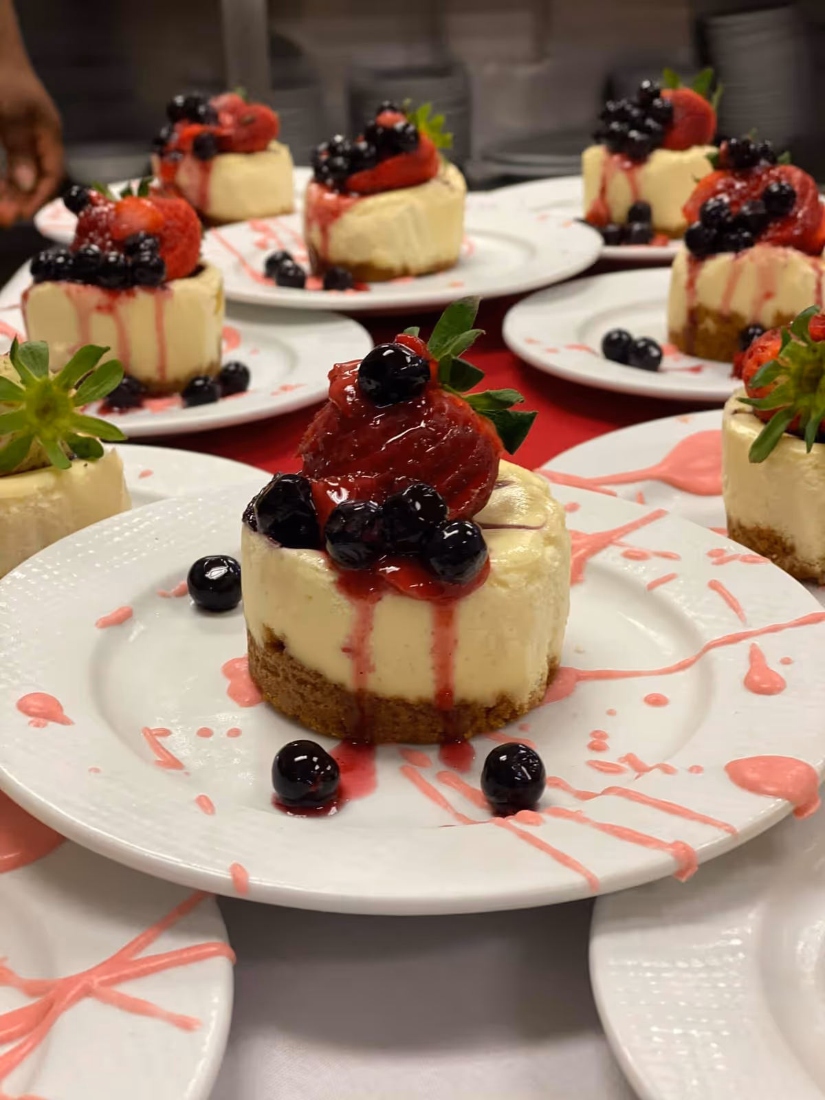 Close-up view of several mini cheesecakes topped with strawberries and blueberries, drizzled with red berry sauce, arranged on white plates.