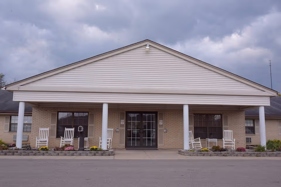 Front exterior view of Crestview Manor Healthcare Center, showing a single-story building with a covered entrance supported by white columns. There are white rocking chairs and small flower beds with colorful flowers on either side of the entrance. The sky is cloudy.