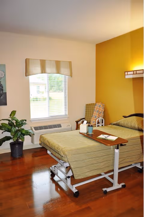 A nursing facility bedroom with a hospital-style bed covered in a greenish-beige bedspread. There is a wooden overbed table with a tissue box, a cup, and a notebook on it. A cushioned chair with a plaid blanket is placed against a mustard yellow accent wall. A window with blinds and a beige valance lets in natural light. A potted plant is on the wooden floor near the window.