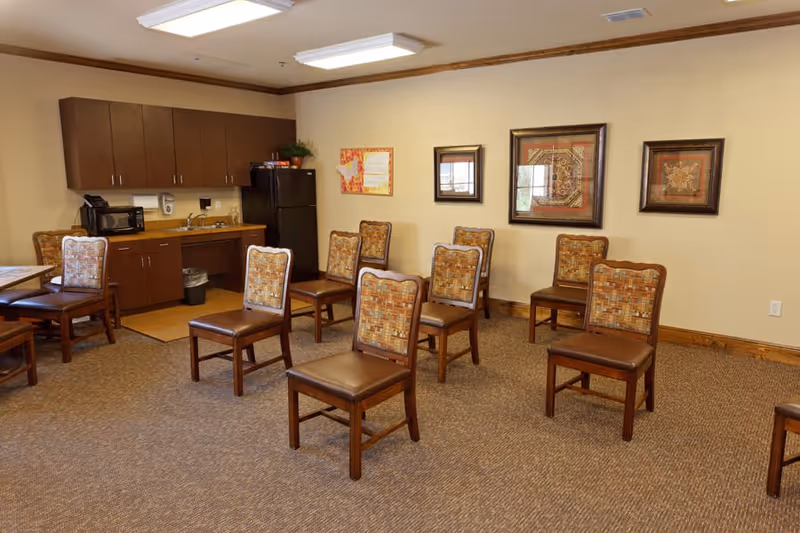 A room with multiple wooden chairs arranged in rows facing forward, a kitchenette area with cabinets, a microwave, a sink, and a refrigerator along one wall, and framed artwork hanging on a beige wall.
