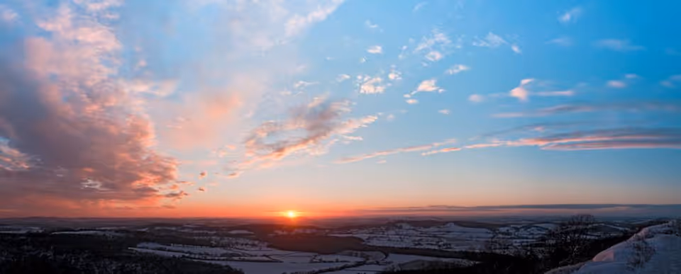 A panoramic view of a snowy landscape at sunset with a colorful sky filled with clouds and the sun near the horizon.